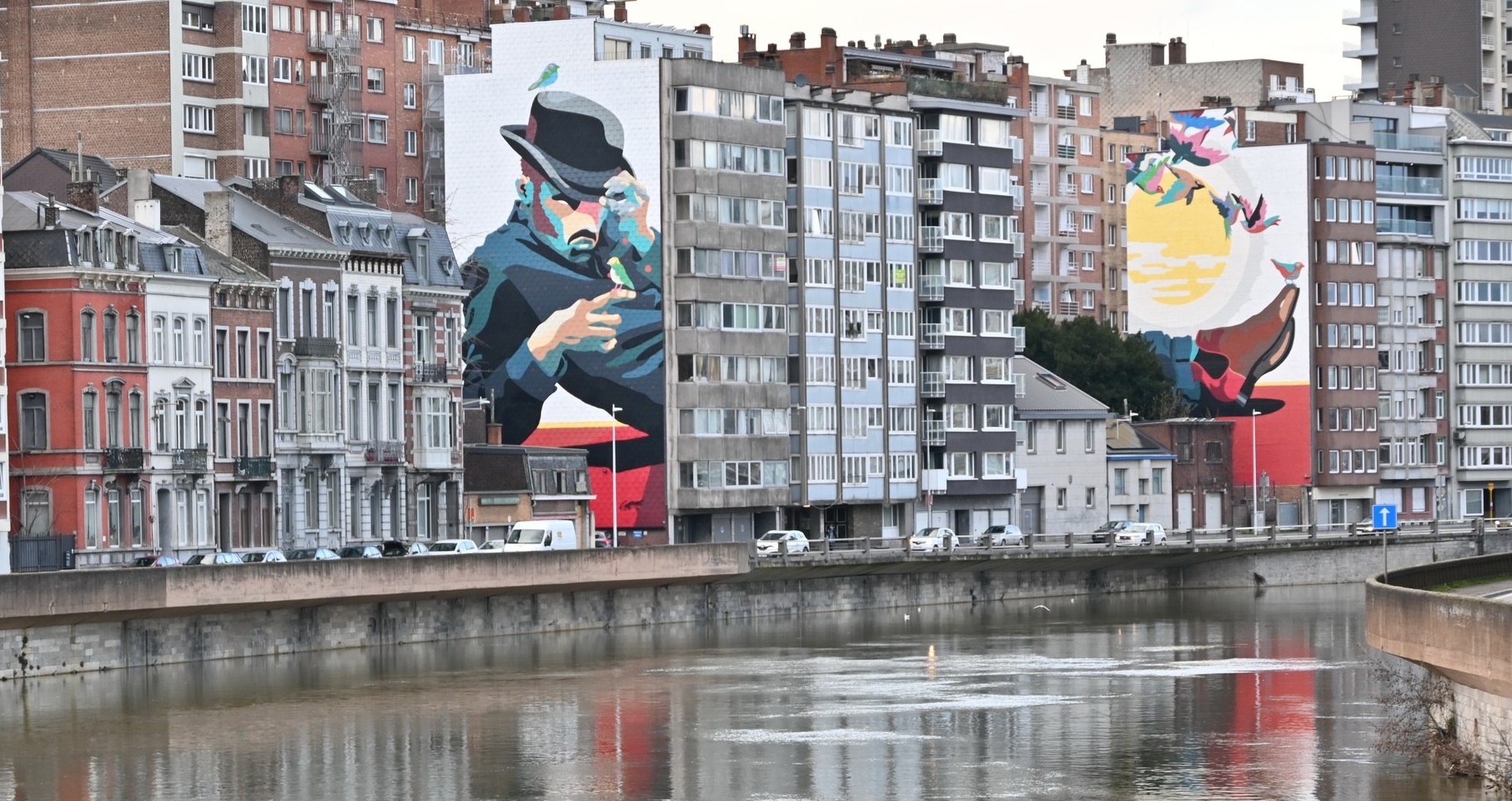 Photo de L'Homme de la Meuse, fresque d'art urbain en diptyque dans le centre-ville de Liège. Homme allongé, posé sur le coude. Les traits de son visage sont géométriques et colorés. Il porte un chapeau.